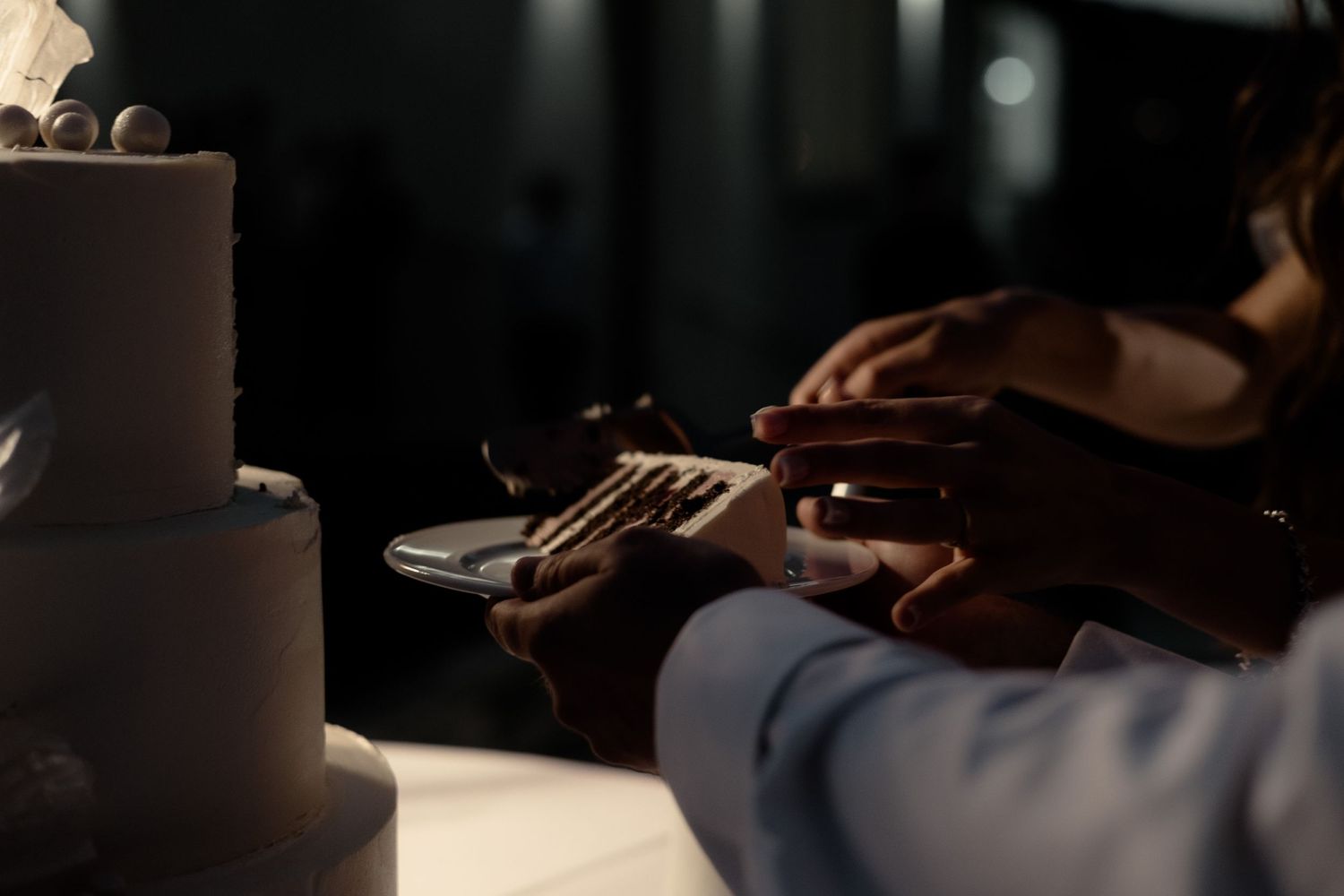 Hands holding a knife to cut into a dark-colored tiered cake in moody lighting.