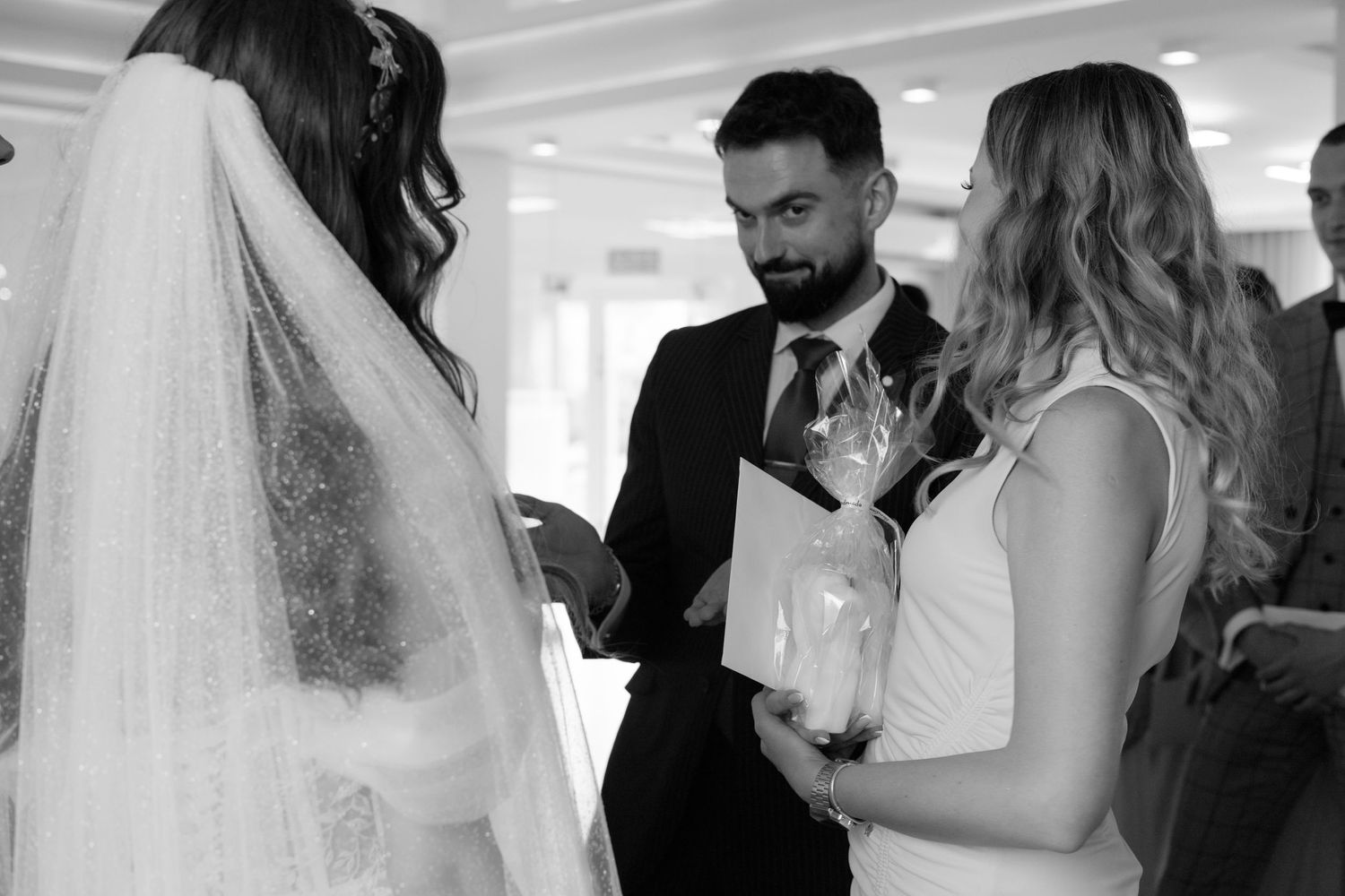 A black and white wedding moment shows guests conversing with the bride who wears a long veil.