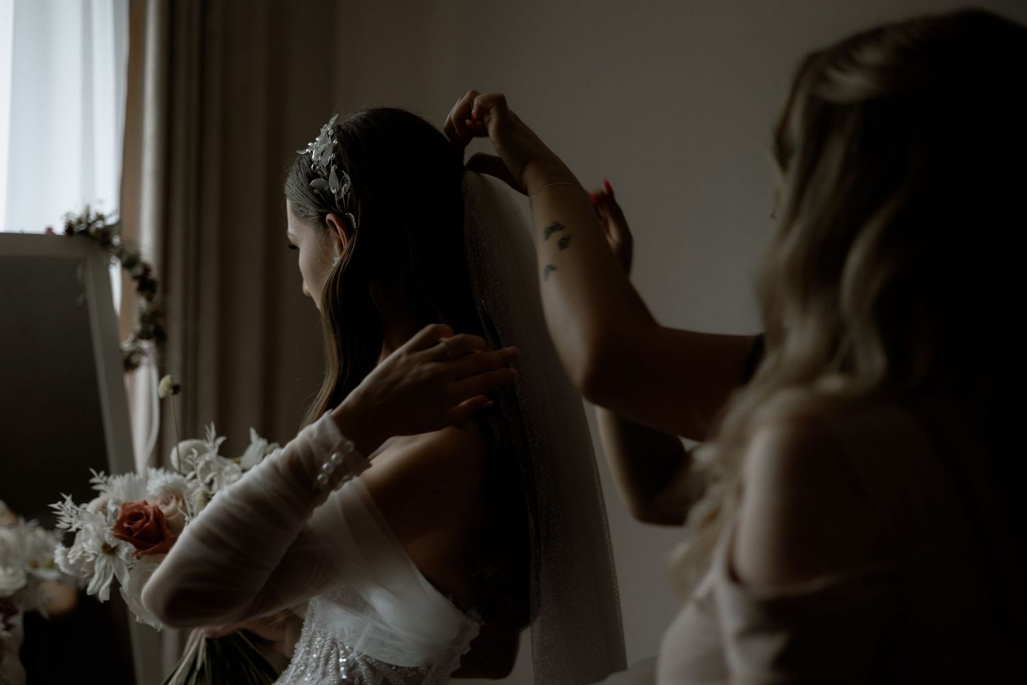 Hands help adjust wedding dress and veil in moody, window-lit getting ready moment.