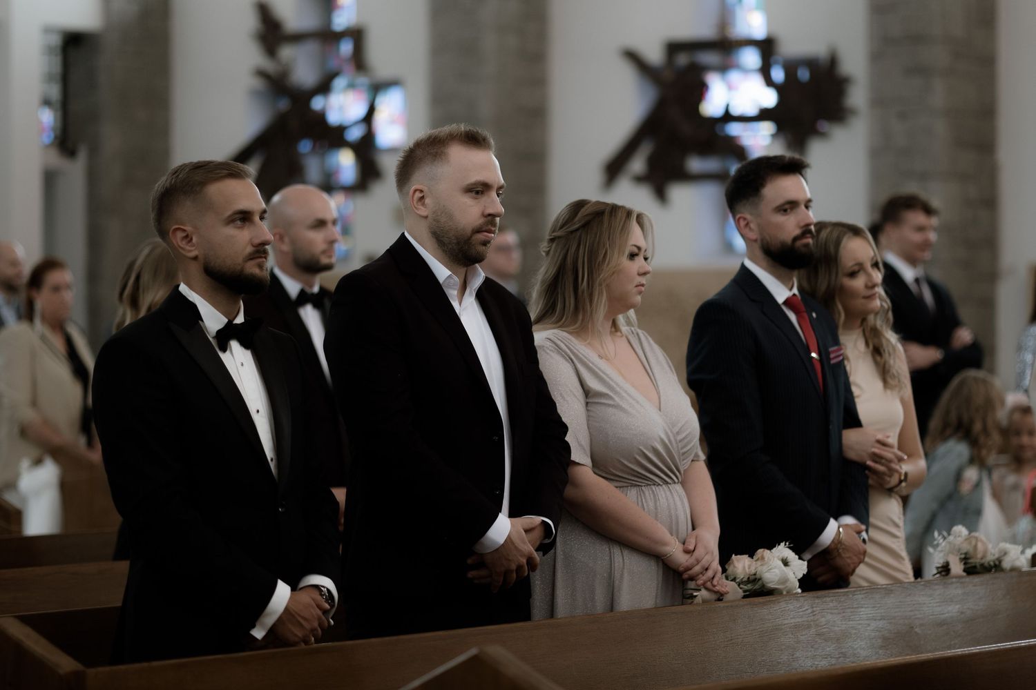 People in formal attire standing in church pews during a wedding ceremony.