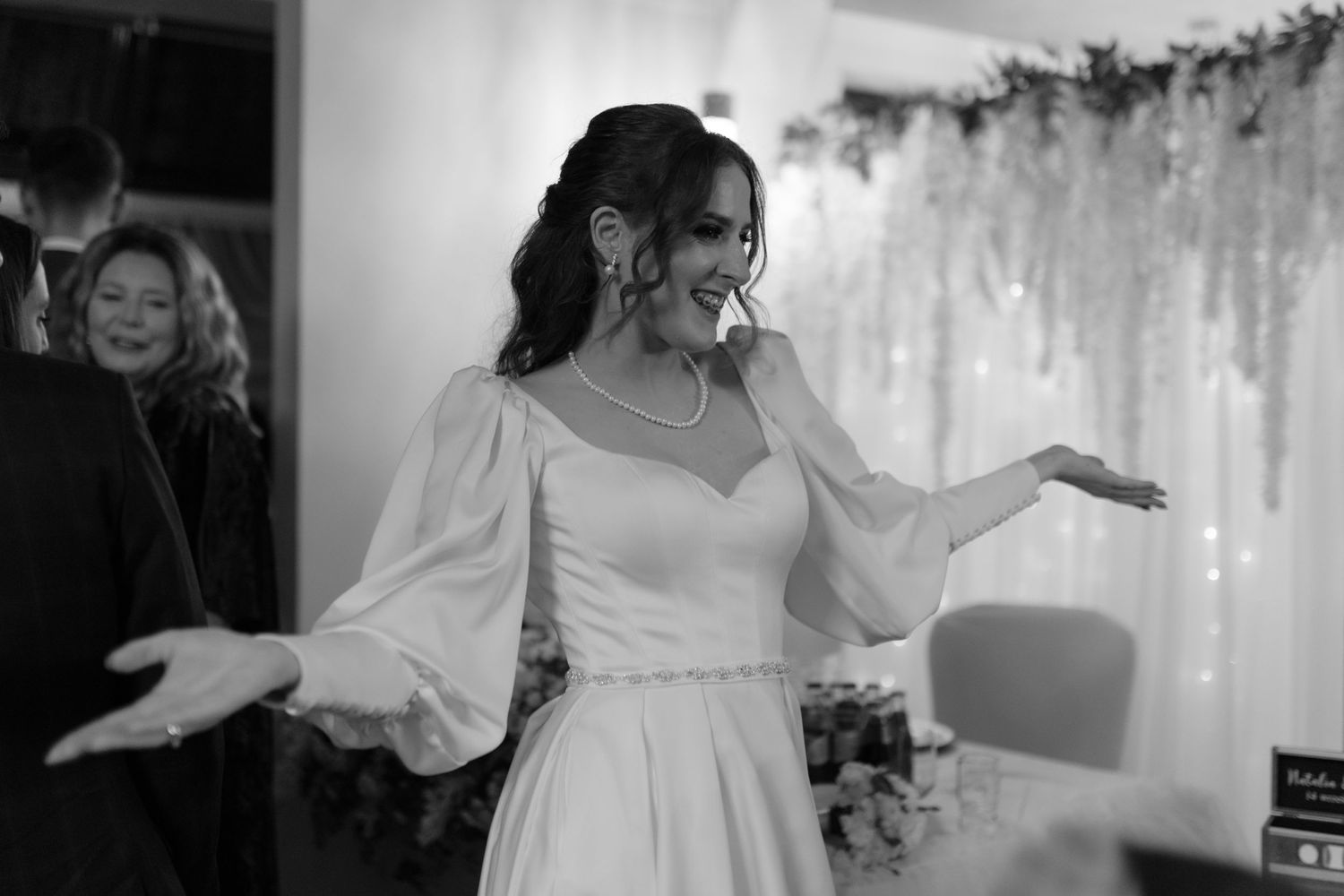 A bride in a long-sleeved wedding gown dances with outstretched arms in a black and white photograph.