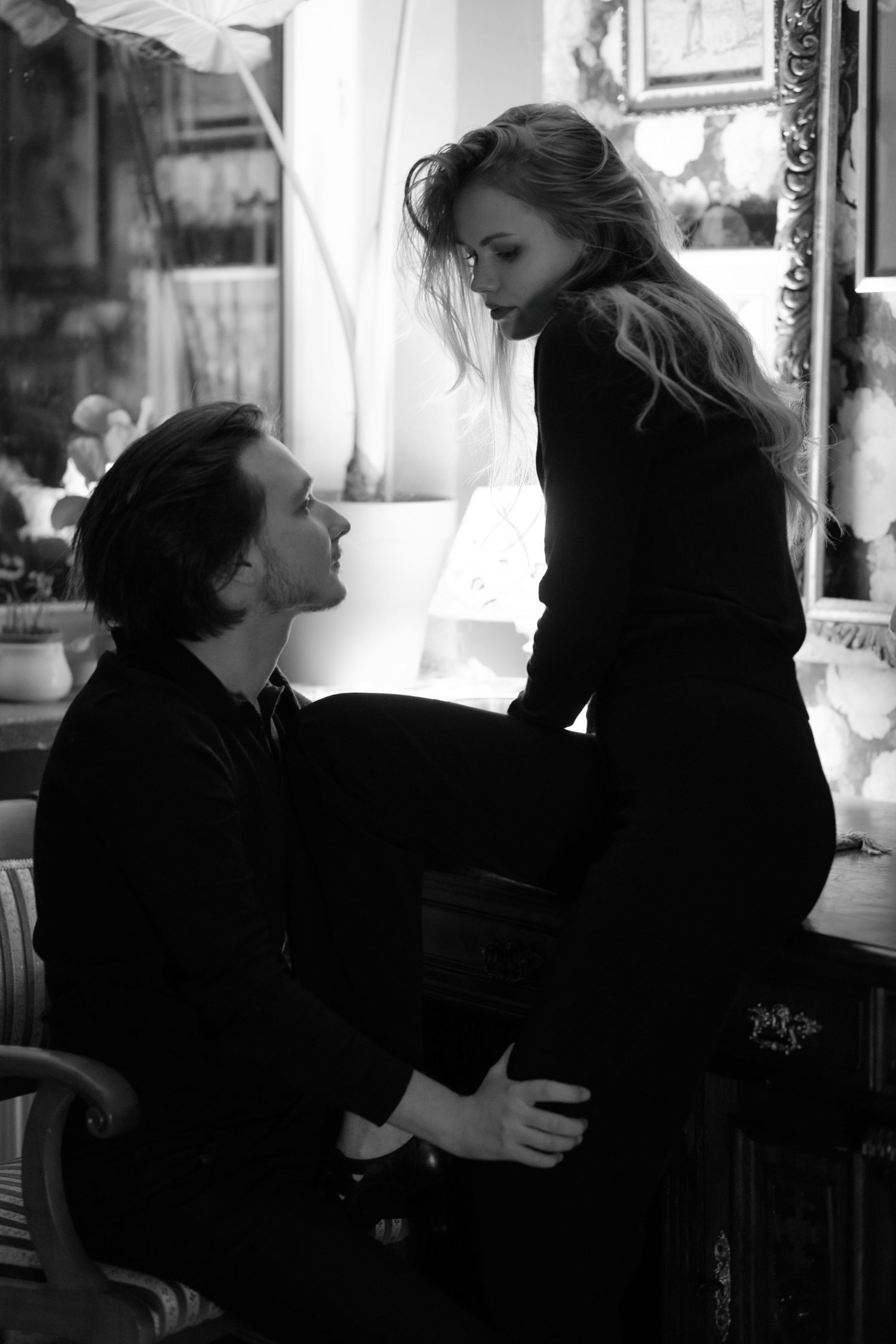 A romantic black and white photograph of a couple in an intimate moment by a window with soft natural lighting.