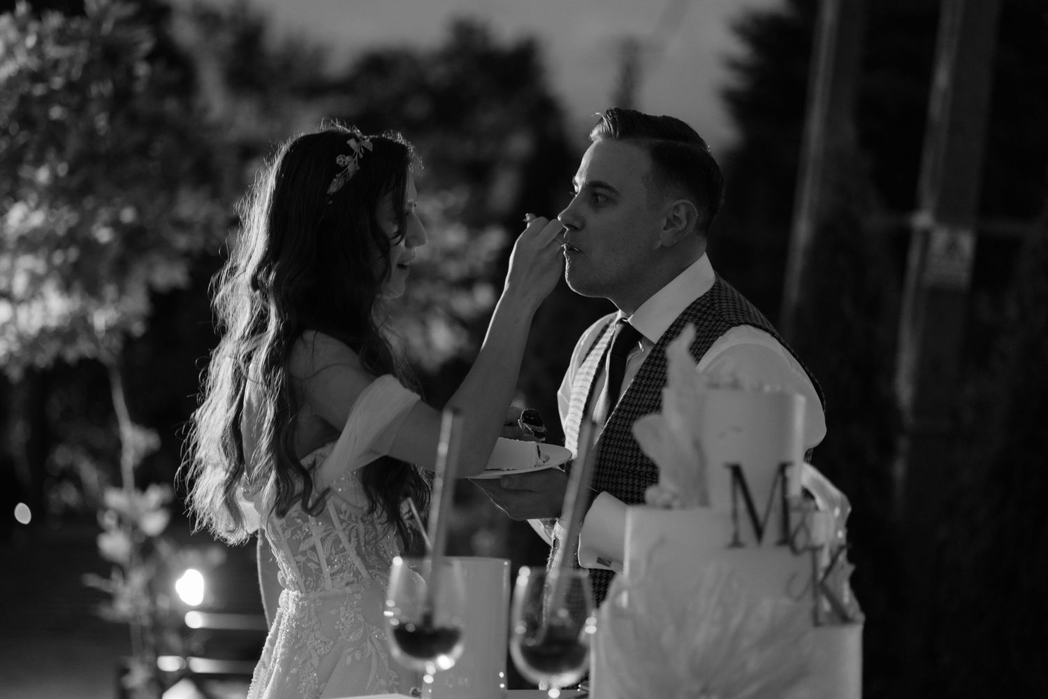 A romantic black and white moment of a couple sharing cake during their outdoor wedding reception.