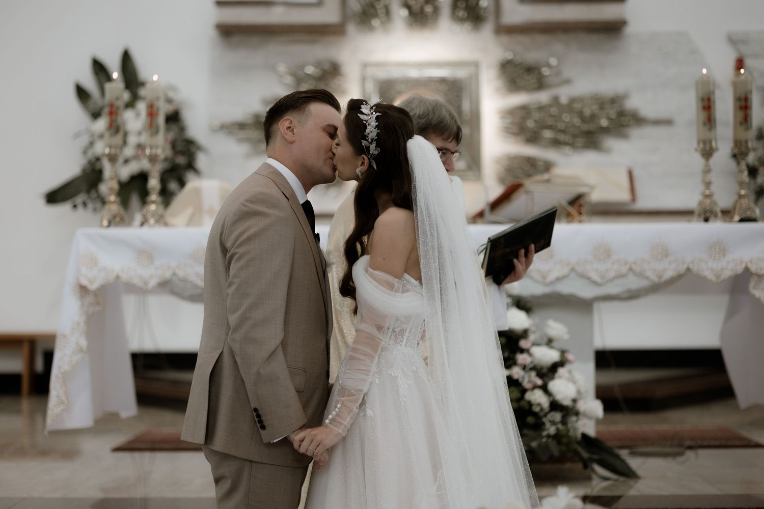 A couple shares a romantic kiss during their wedding ceremony in an elegant church decorated with white floral arrangements.