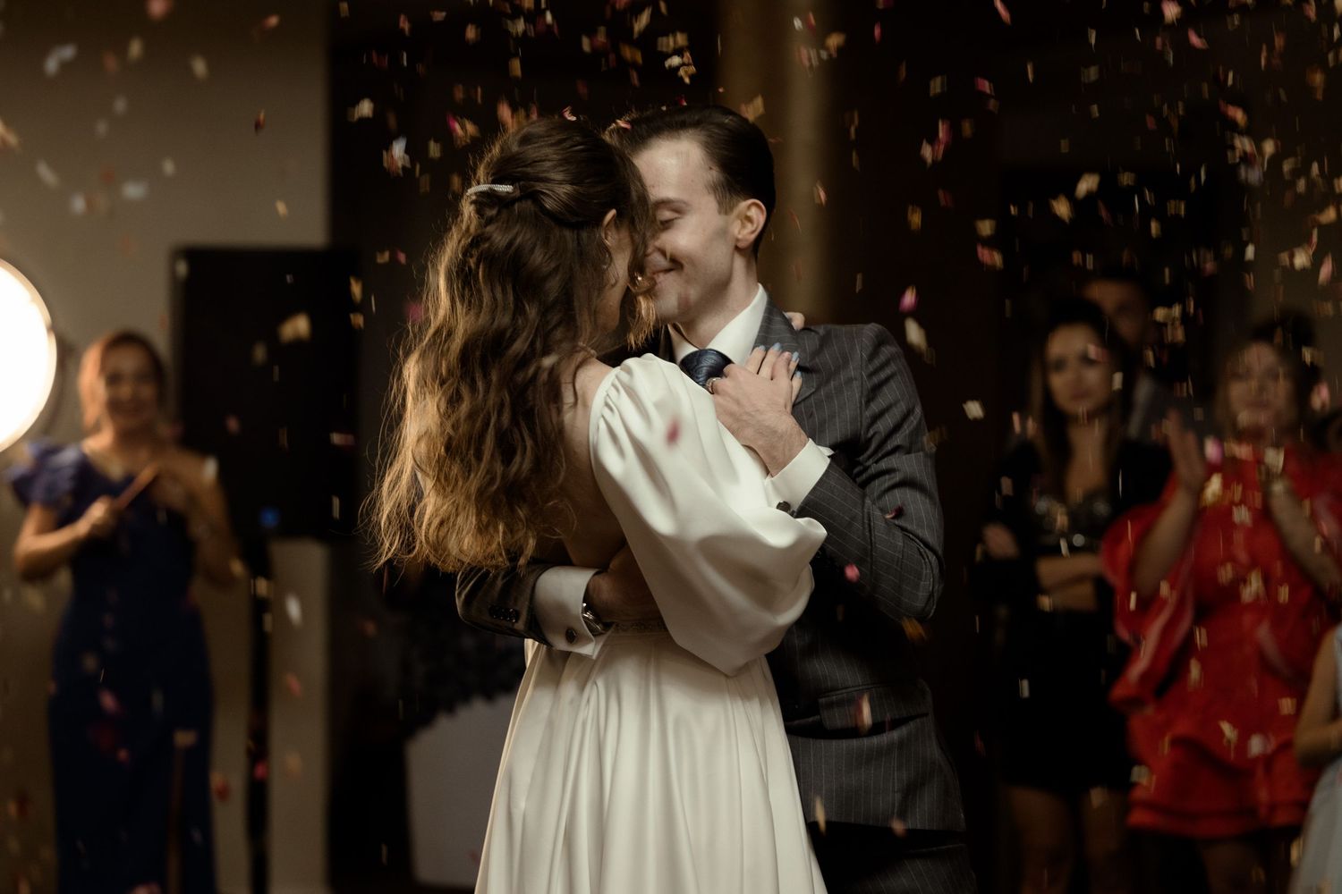 A couple shares their first dance at their wedding celebration while confetti falls around them in a dimly lit room.