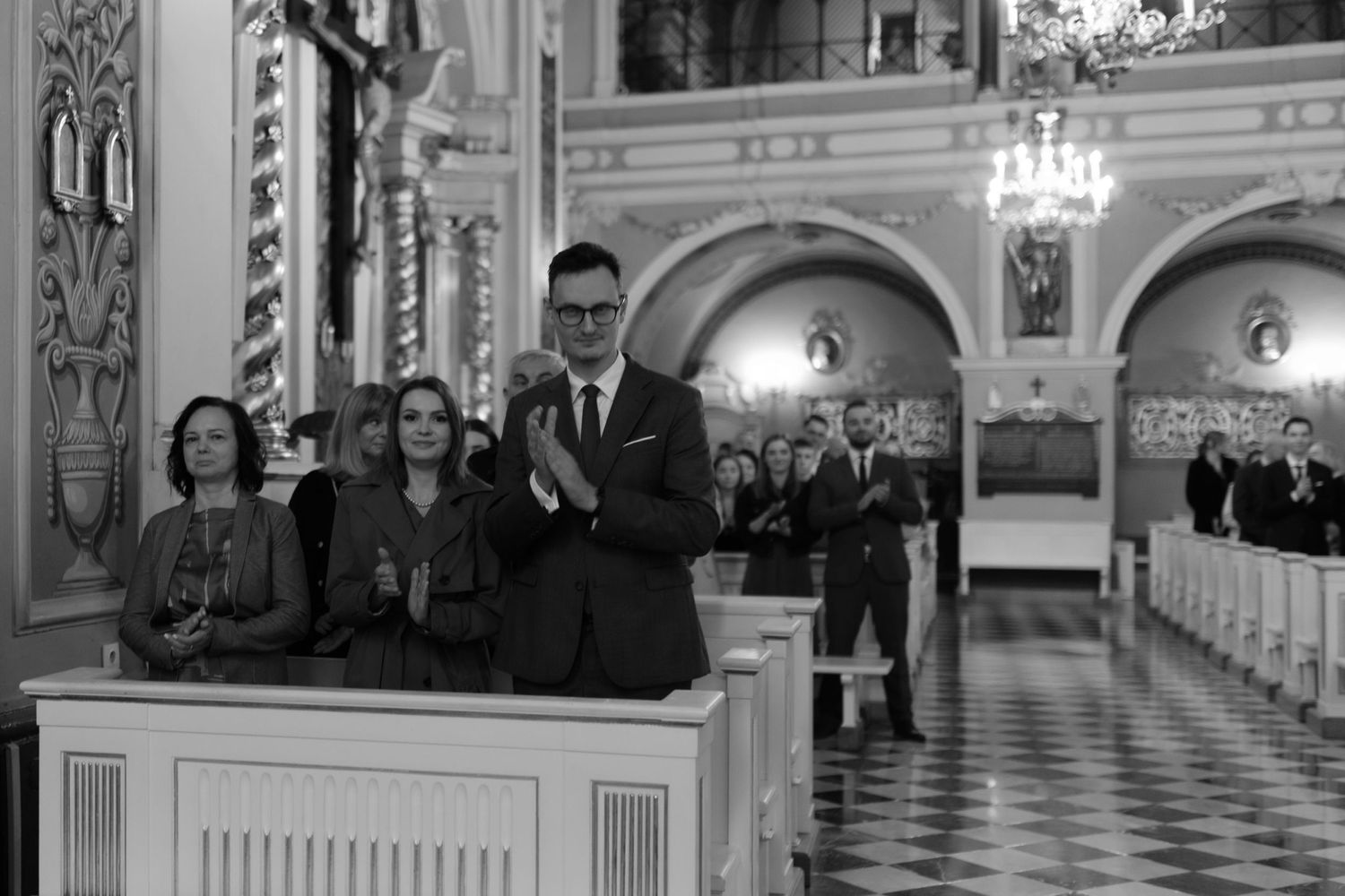 A group stands in a traditional church balcony with ornate architecture and chandeliers visible in the background.