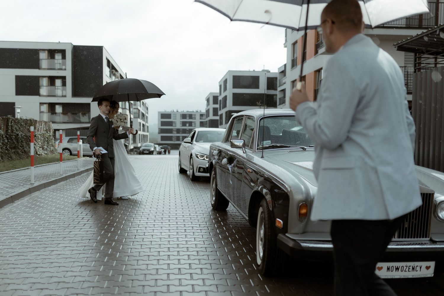 A vintage Rolls-Royce parked on a wet cobblestone street while people with umbrellas take shelter from the rain.