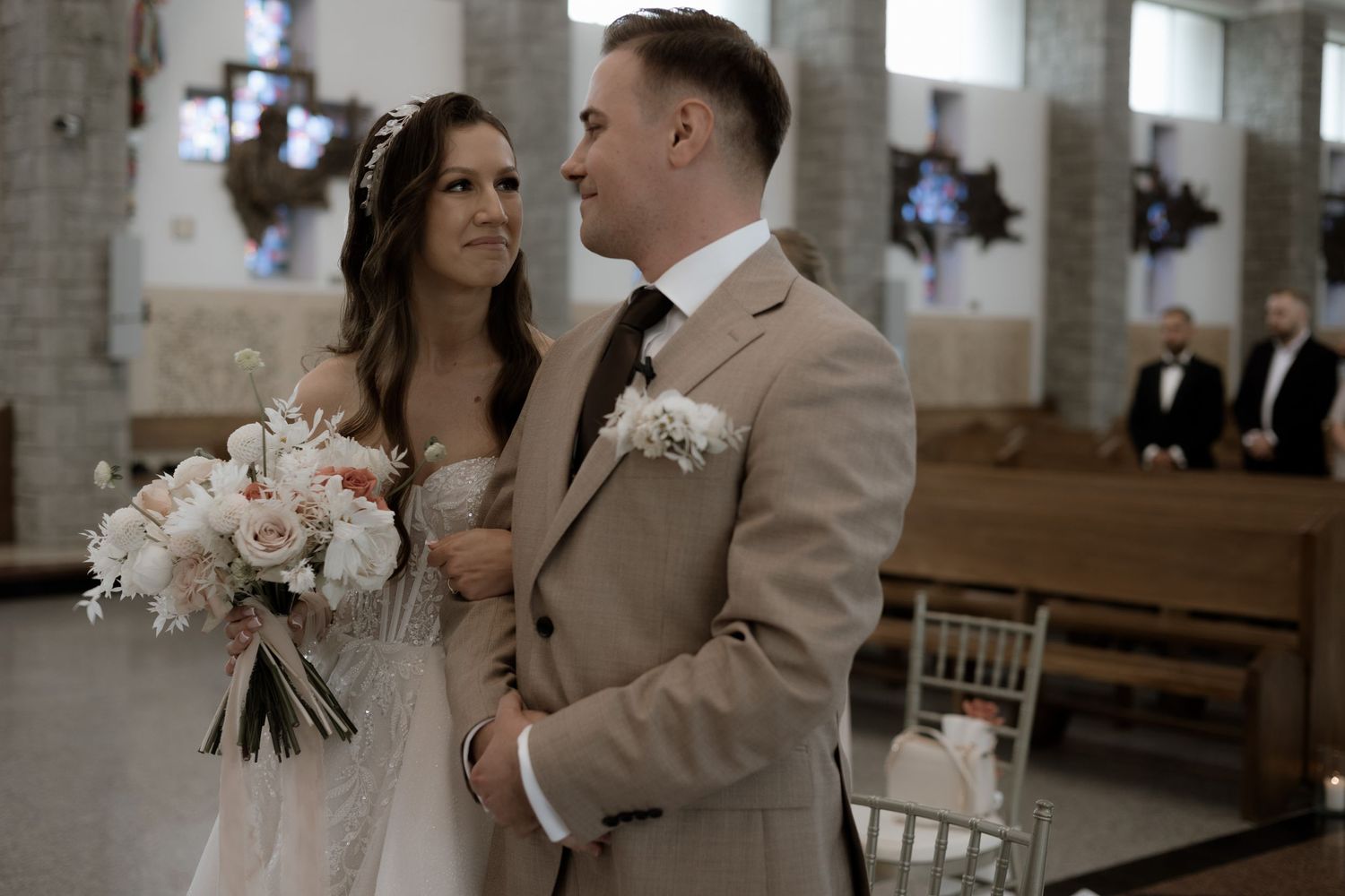Bride and groom share an intimate moment in a church with blush floral bouquet and tan suits.
