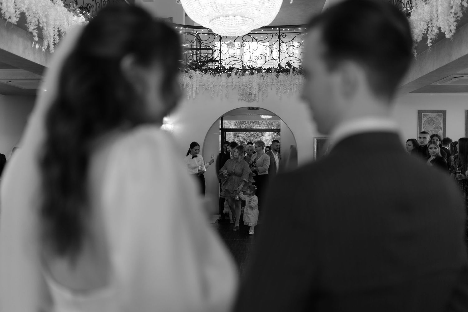 A black and white photo of a couple during a wedding ceremony at an ornate indoor venue, shot from behind.