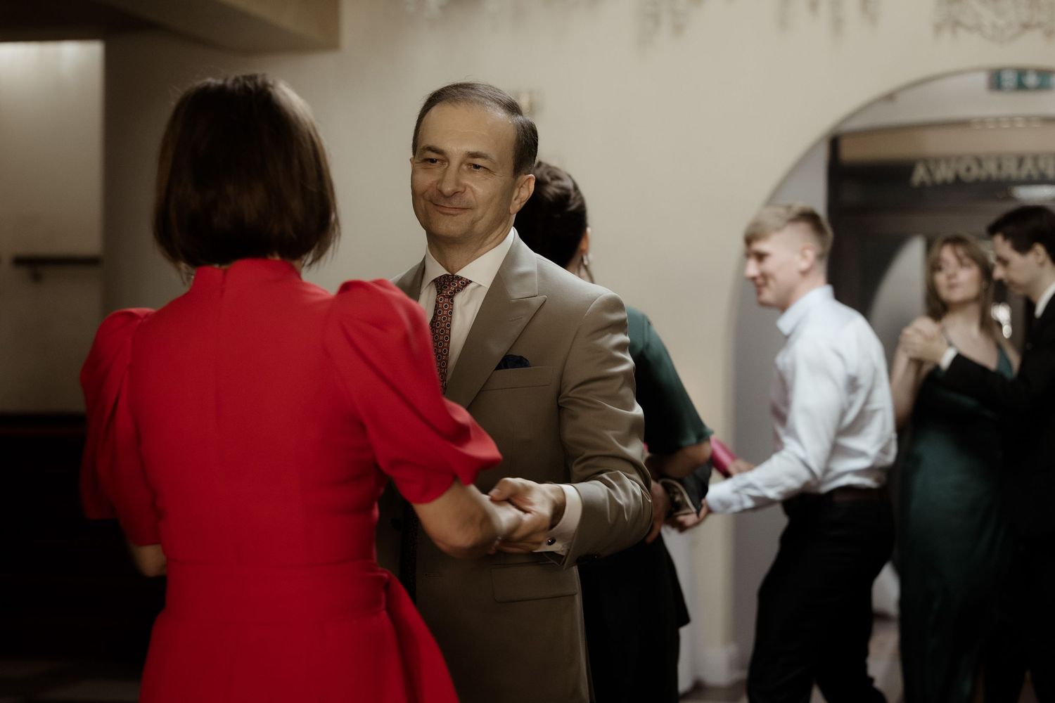 A couple dances together at a social gathering, with the woman wearing red and the man in a brown suit.