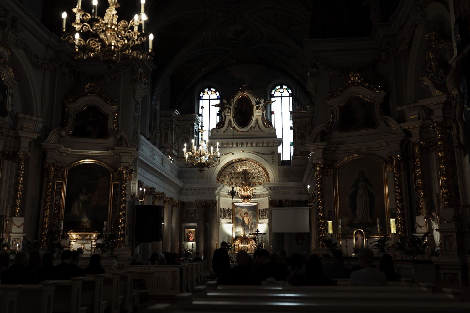 Interior view of an ornate baroque church with chandelier, arches and gilded decorations in dark atmospheric lighting.