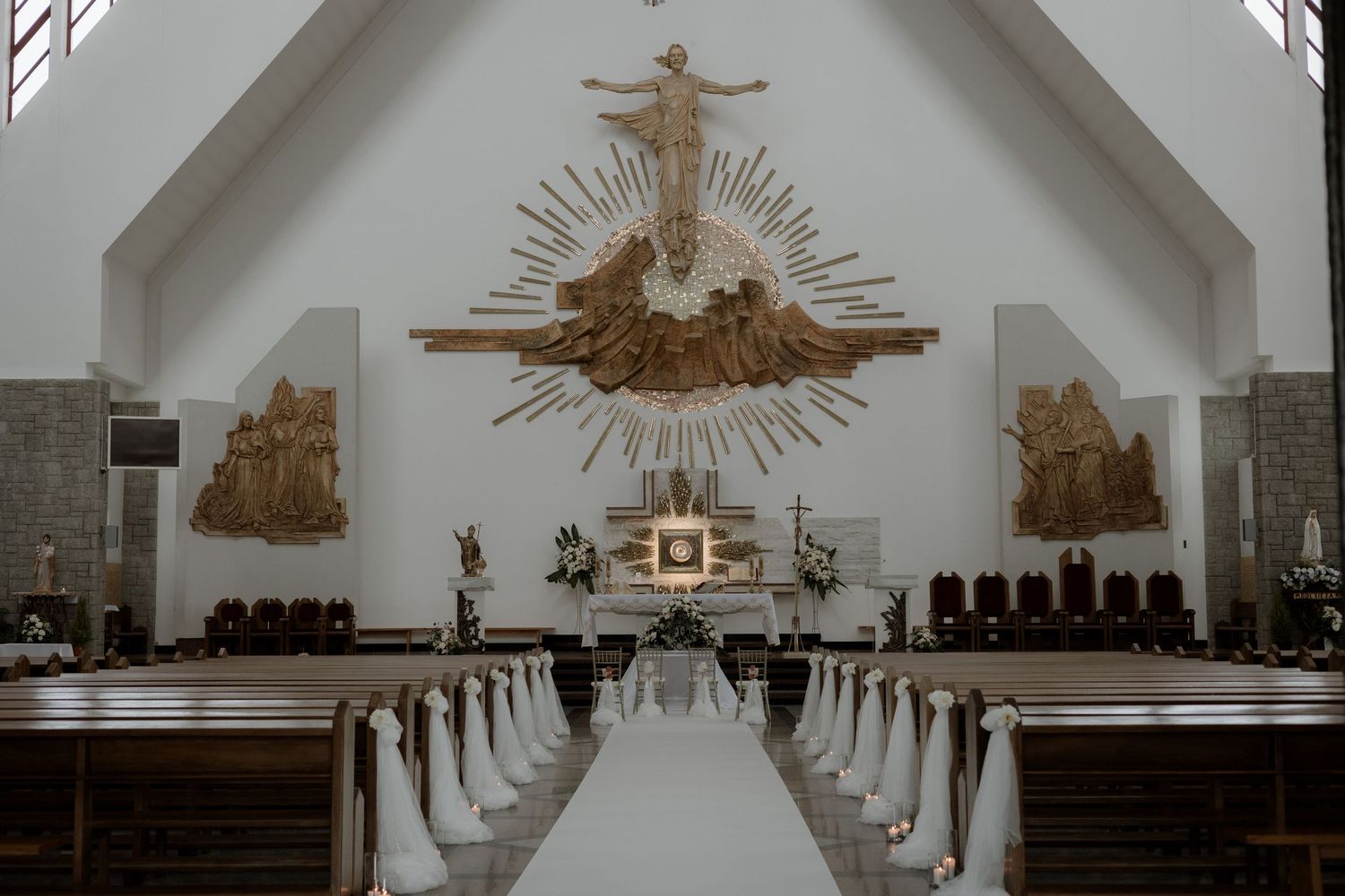 Interior view of a modern Catholic church with white walls, wooden pews, and a large crucifix mounted above the altar.