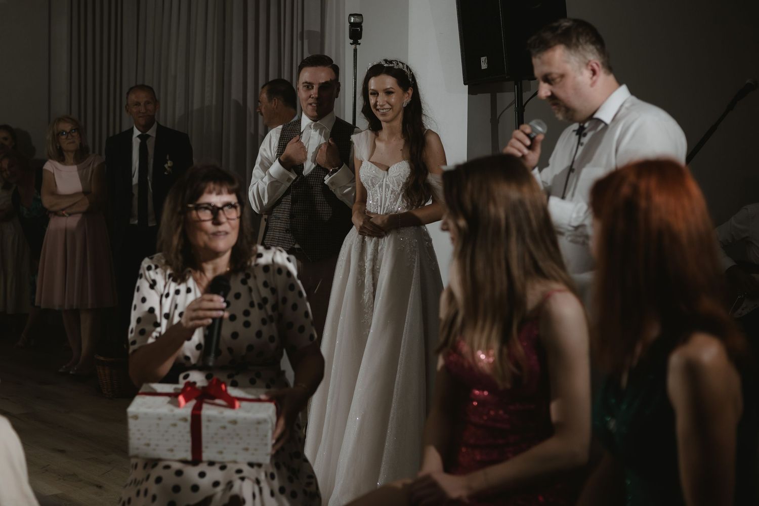 Wedding reception guests and formally dressed attendees gathered at an indoor celebration with a present being opened.