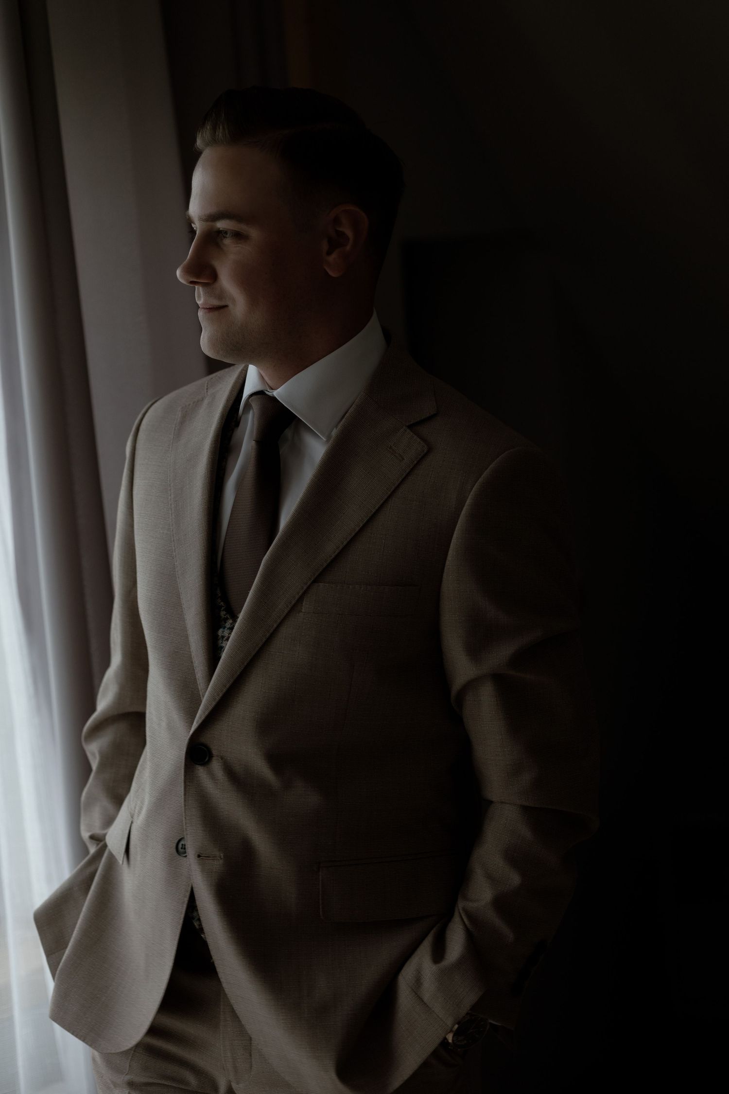 A person in a beige suit and dark tie stands in moody lighting near a window looking to the side.