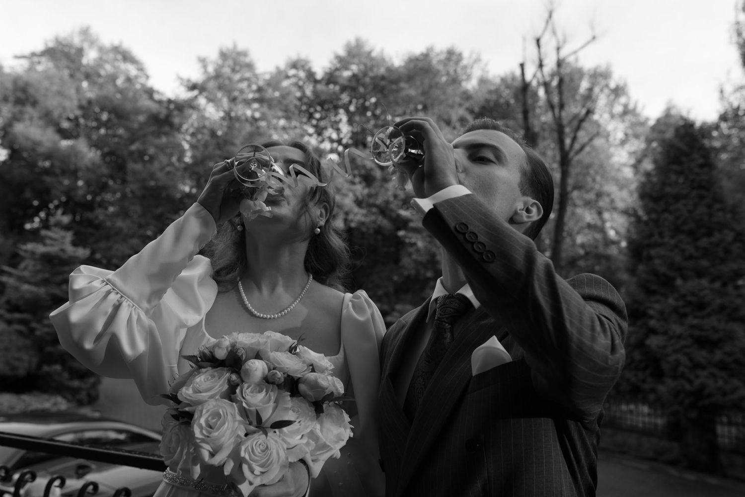 A black and white photograph of a bride and groom blowing flower petals into the air during an outdoor celebration.