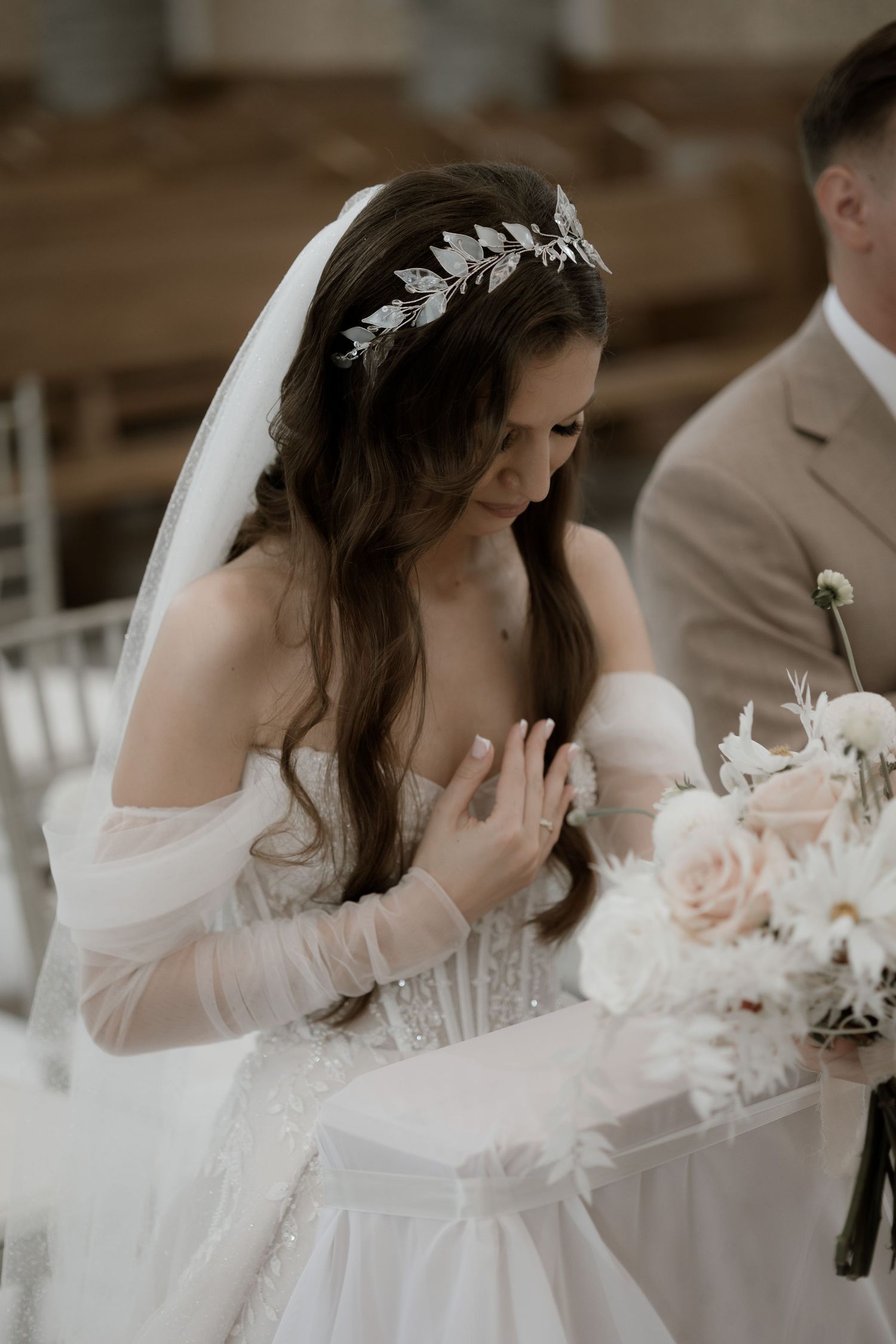 Bride in elegant off-shoulder wedding gown with delicate leaf headpiece sits at reception table surrounded by blush roses.