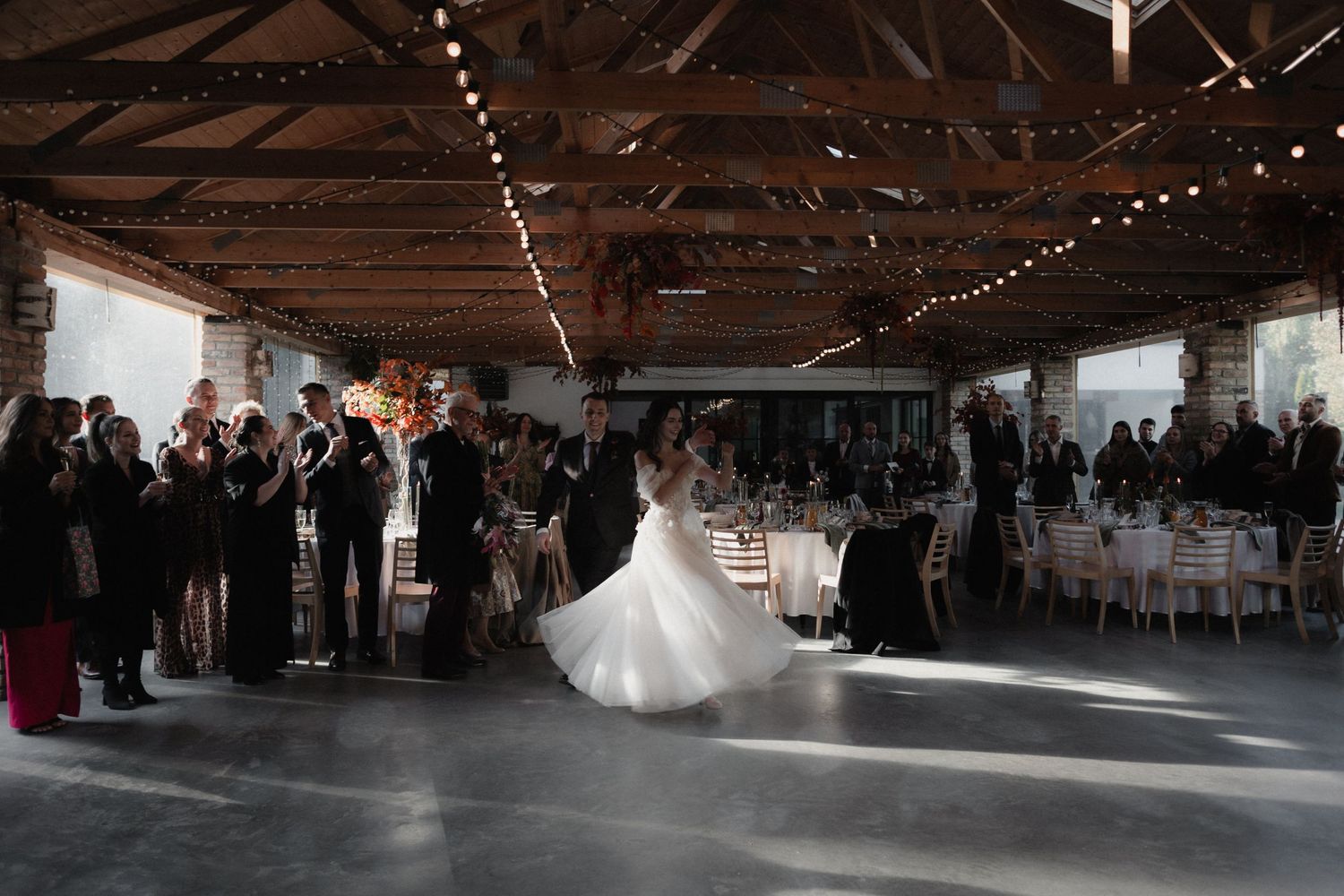 Wedding guests dance on a rustic barn floor under twinkling string lights during a reception celebration.