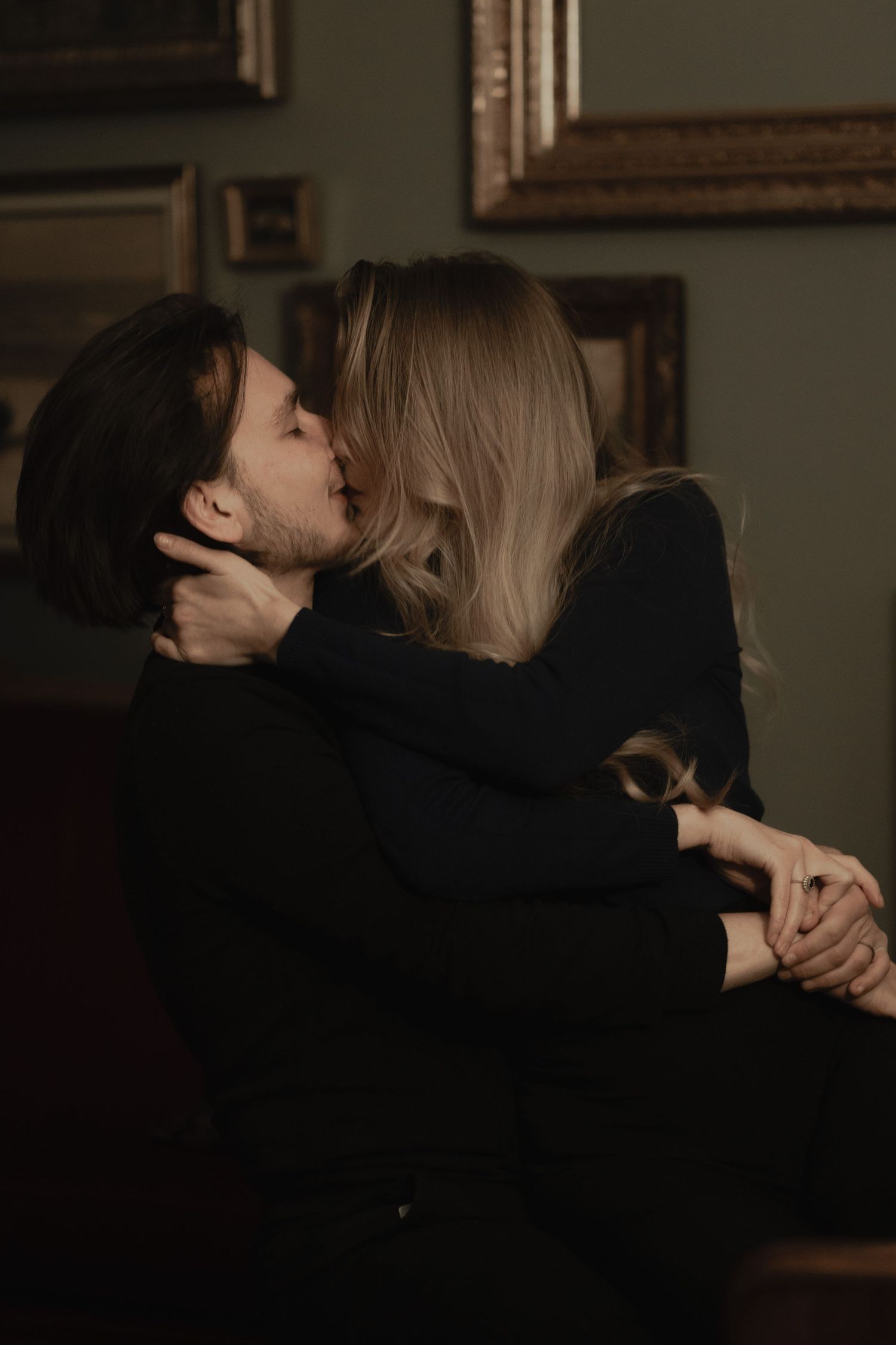 A romantic couple embraces in dark clothing while sharing an intimate kiss in a dimly lit room with framed art.