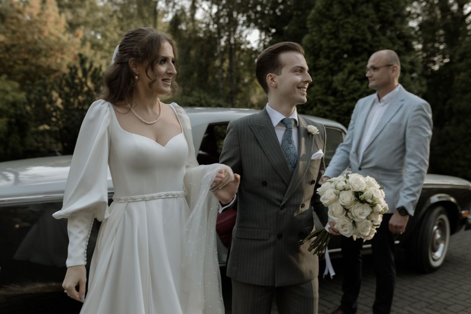 A wedding couple shares a joyful moment together while holding a white bouquet near a car outdoors.