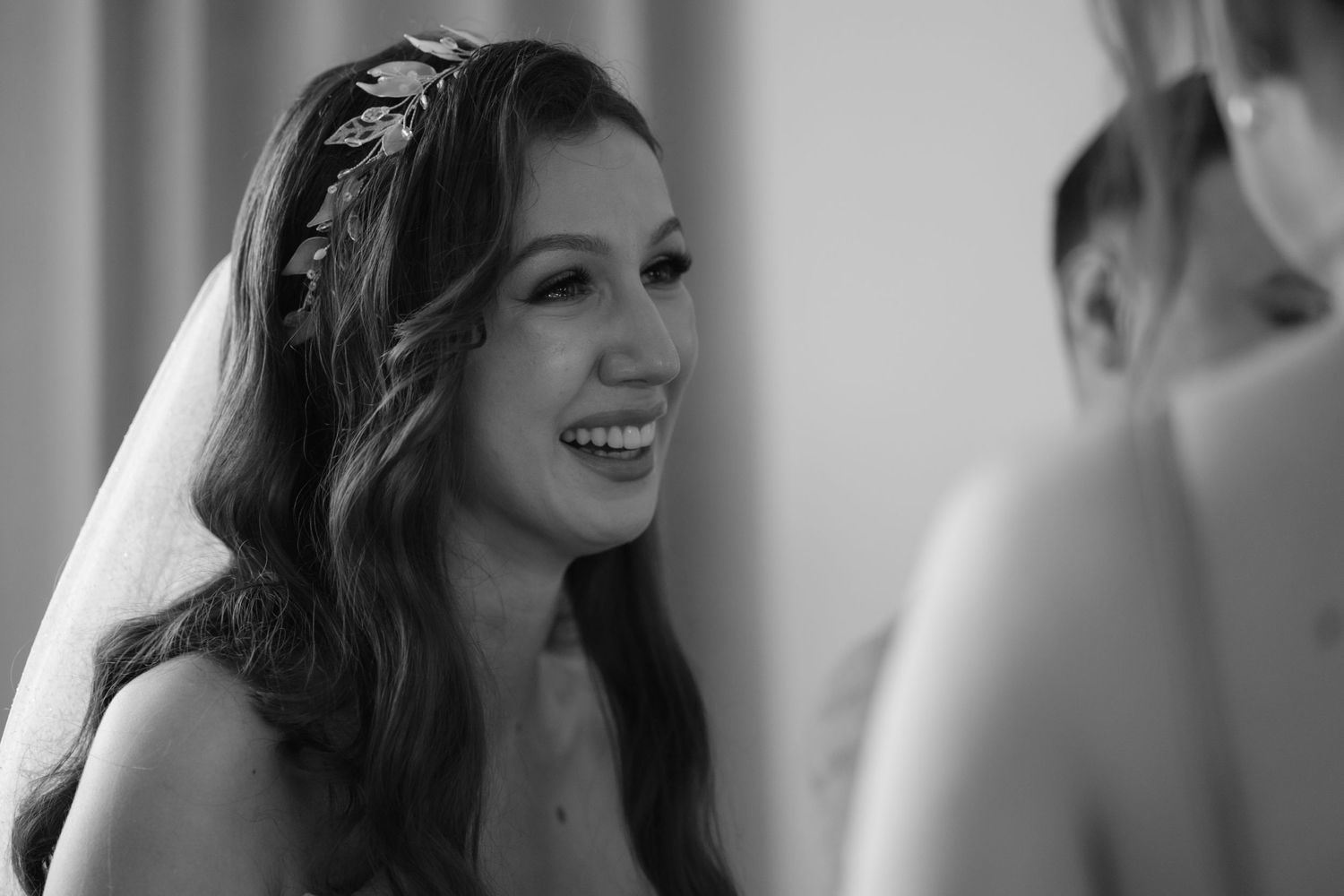 Black and white photo of a bride smiling during her wedding preparations, wearing a delicate veil and floral headpiece.