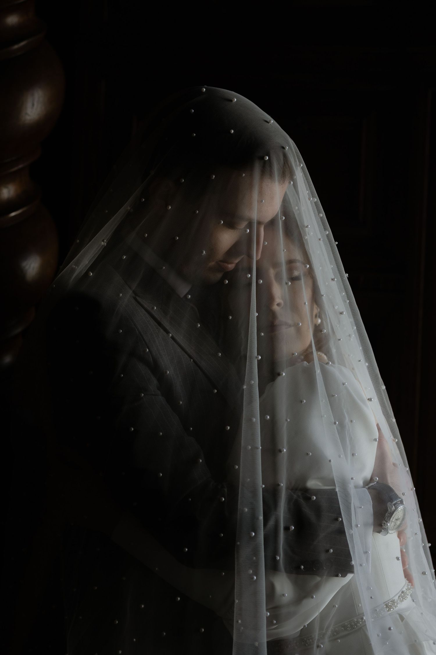 Moody artistic portrait through delicate pearl-dotted sheer white bridal veil in dark atmospheric lighting.
