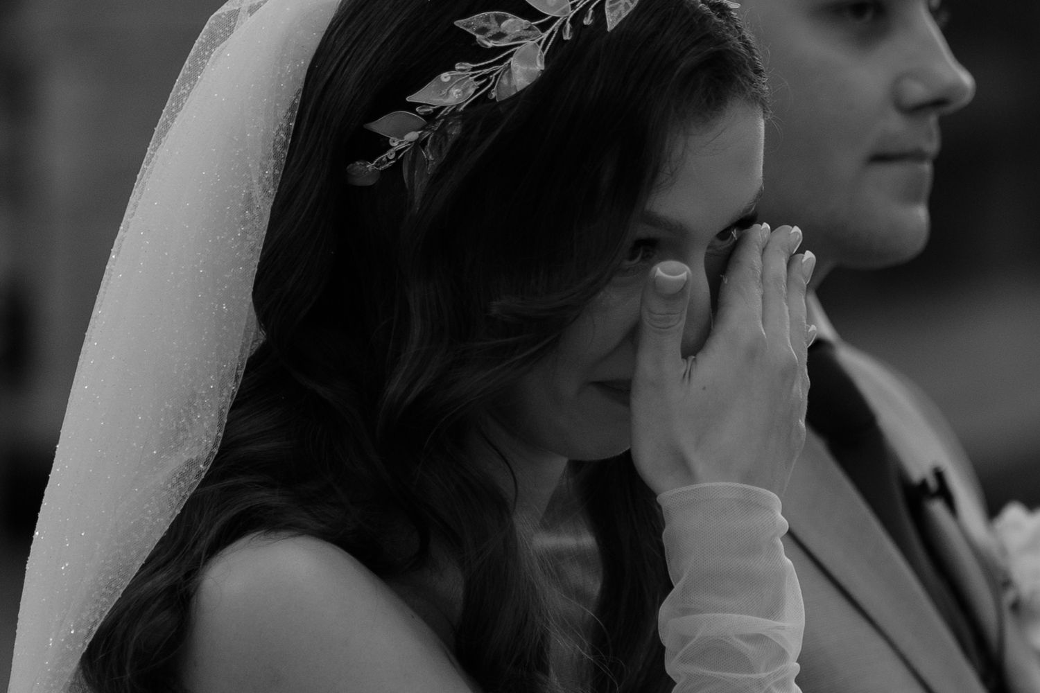 A bride in a white veil and floral crown wipes away tears during an emotional wedding ceremony moment.