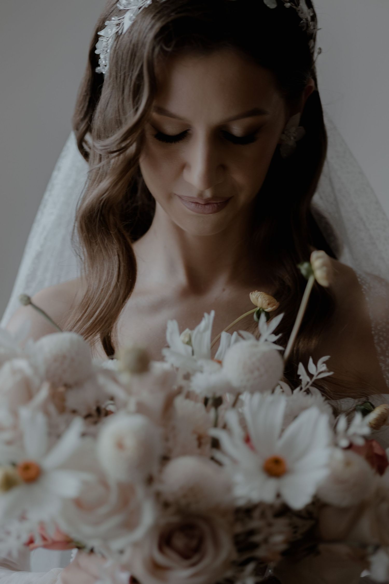A bride in white veil looks down while holding a soft pastel bouquet of roses and daisies in a moody portrait.