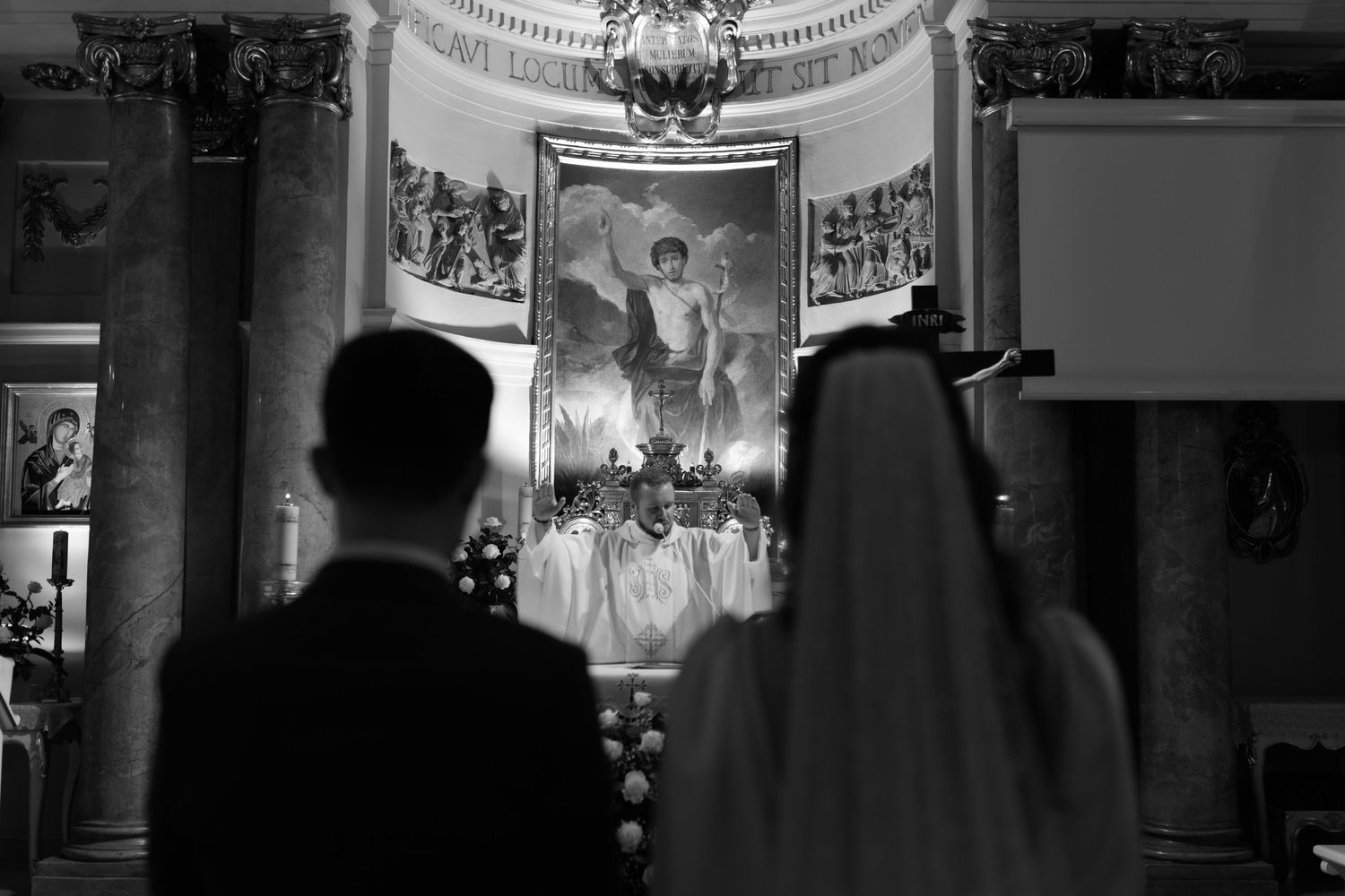 A silhouetted couple watches a religious ceremony at a church altar with ornate architectural details in black and white.
