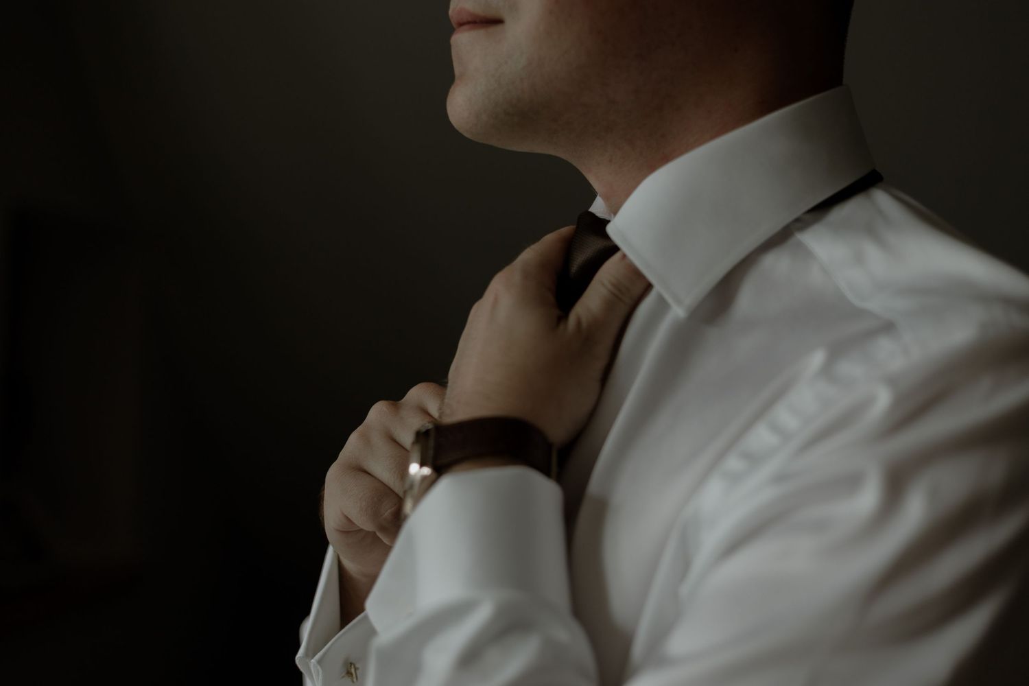 A person in a crisp white dress shirt adjusts their dark necktie against a moody, shadowed background.