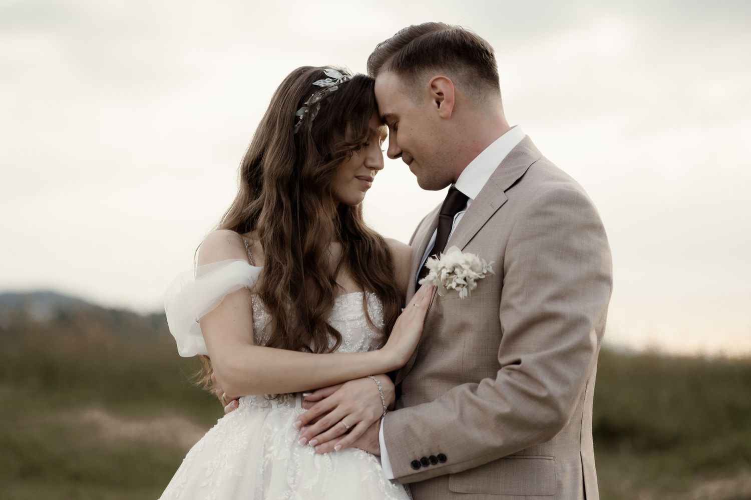 A romantic wedding couple embraces in a dreamy outdoor setting at sunset wearing a classic white dress and tan suit.