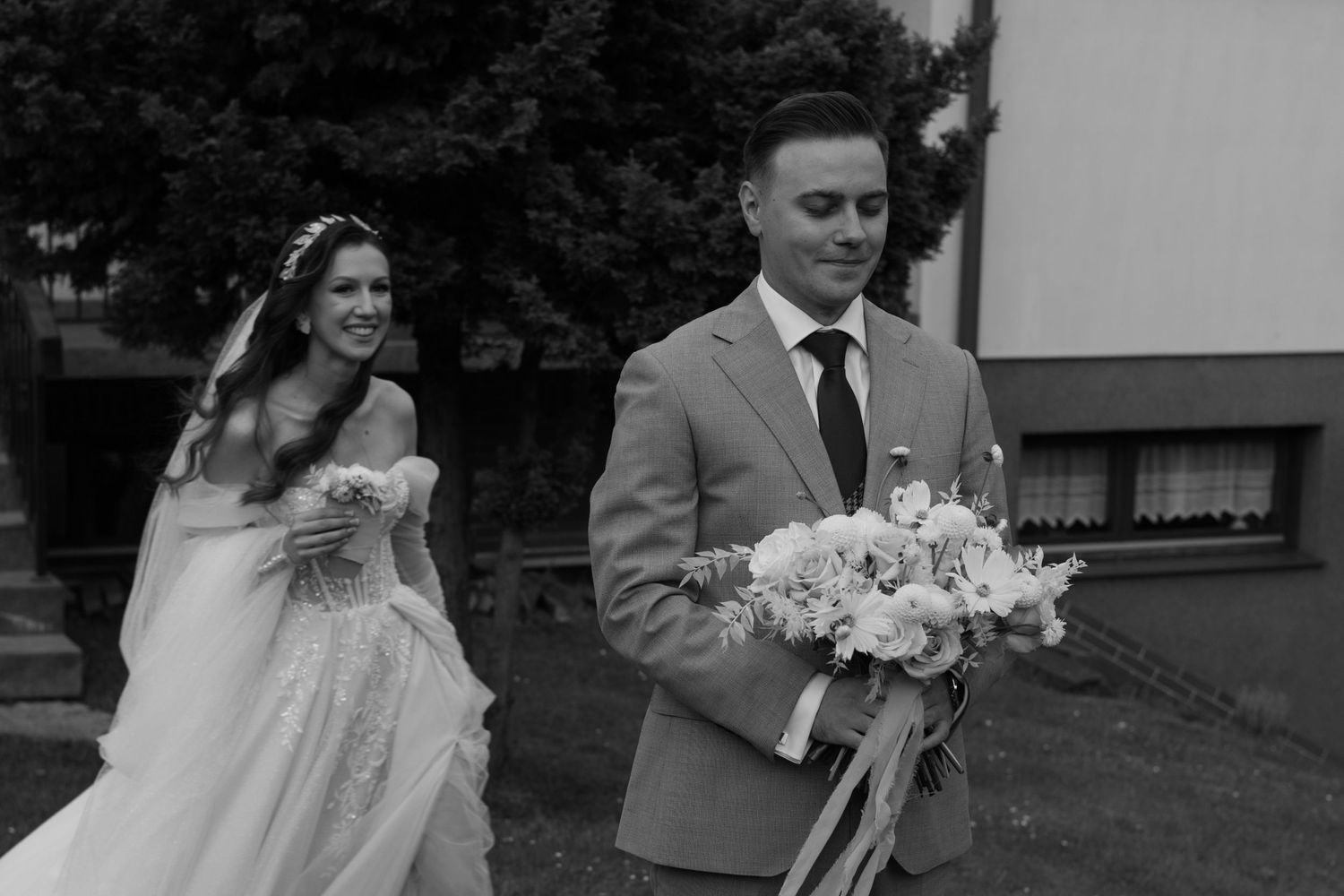 A black and white photograph of newlyweds walking together outdoors, with the bride in a flowing gown and carrying flowers.