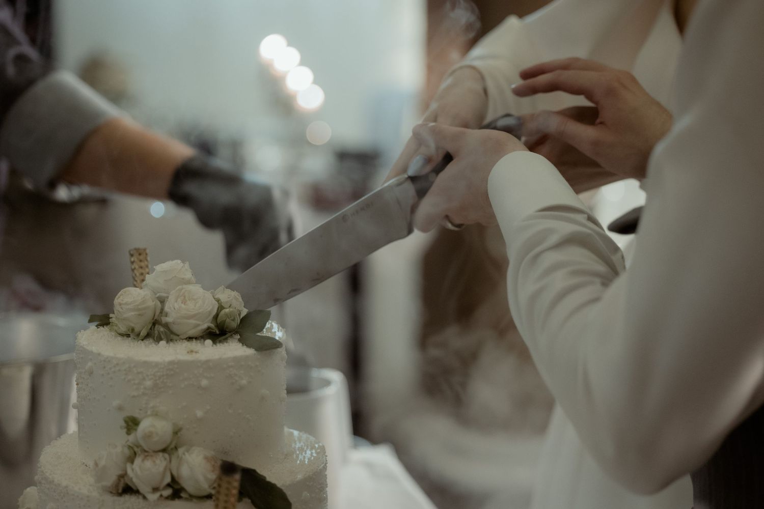 A close-up view of hands cutting into a white tiered wedding cake during a reception.