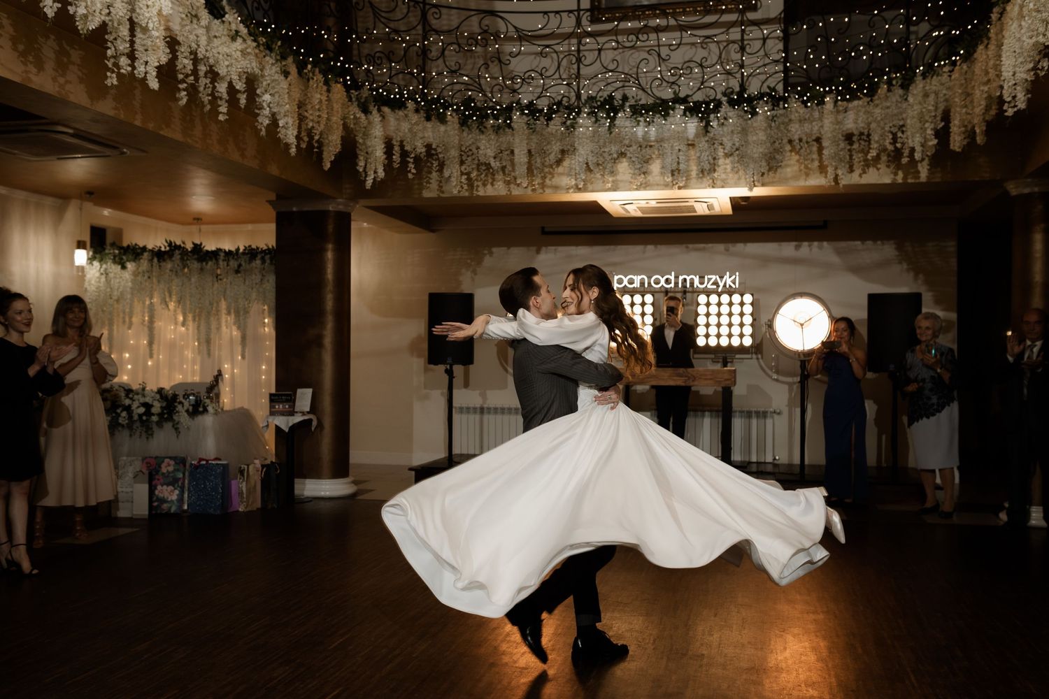 A couple performs a romantic twirling dance move under hanging lights at an indoor wedding reception venue.
