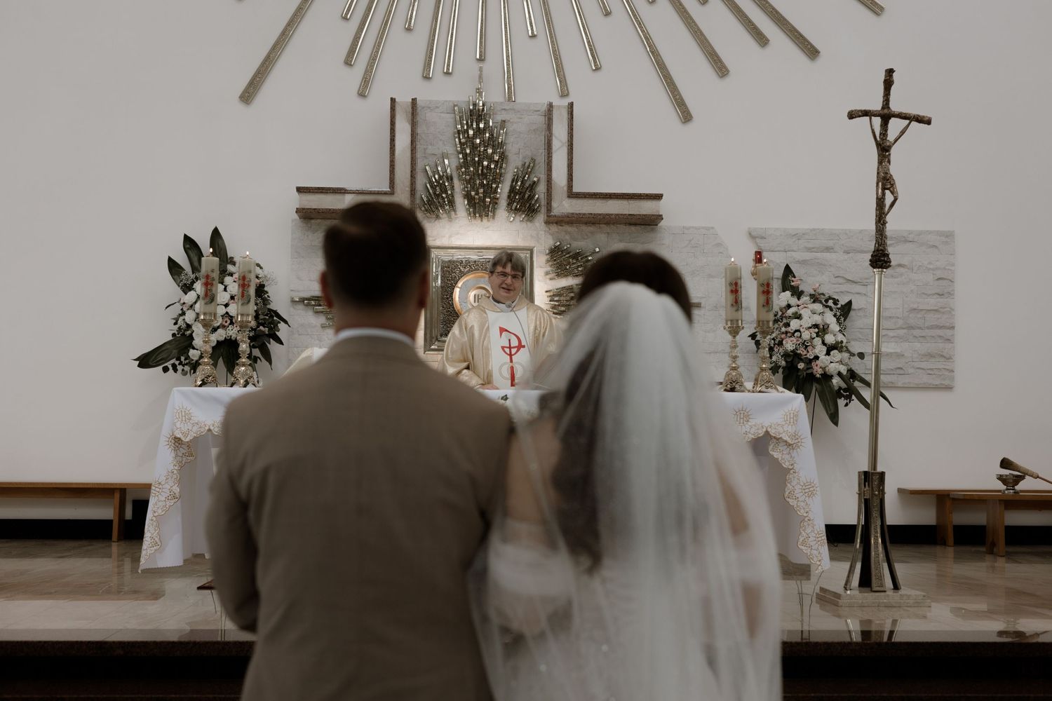 A couple kneels at the altar during their wedding ceremony with white floral arrangements and a crucifix on display.