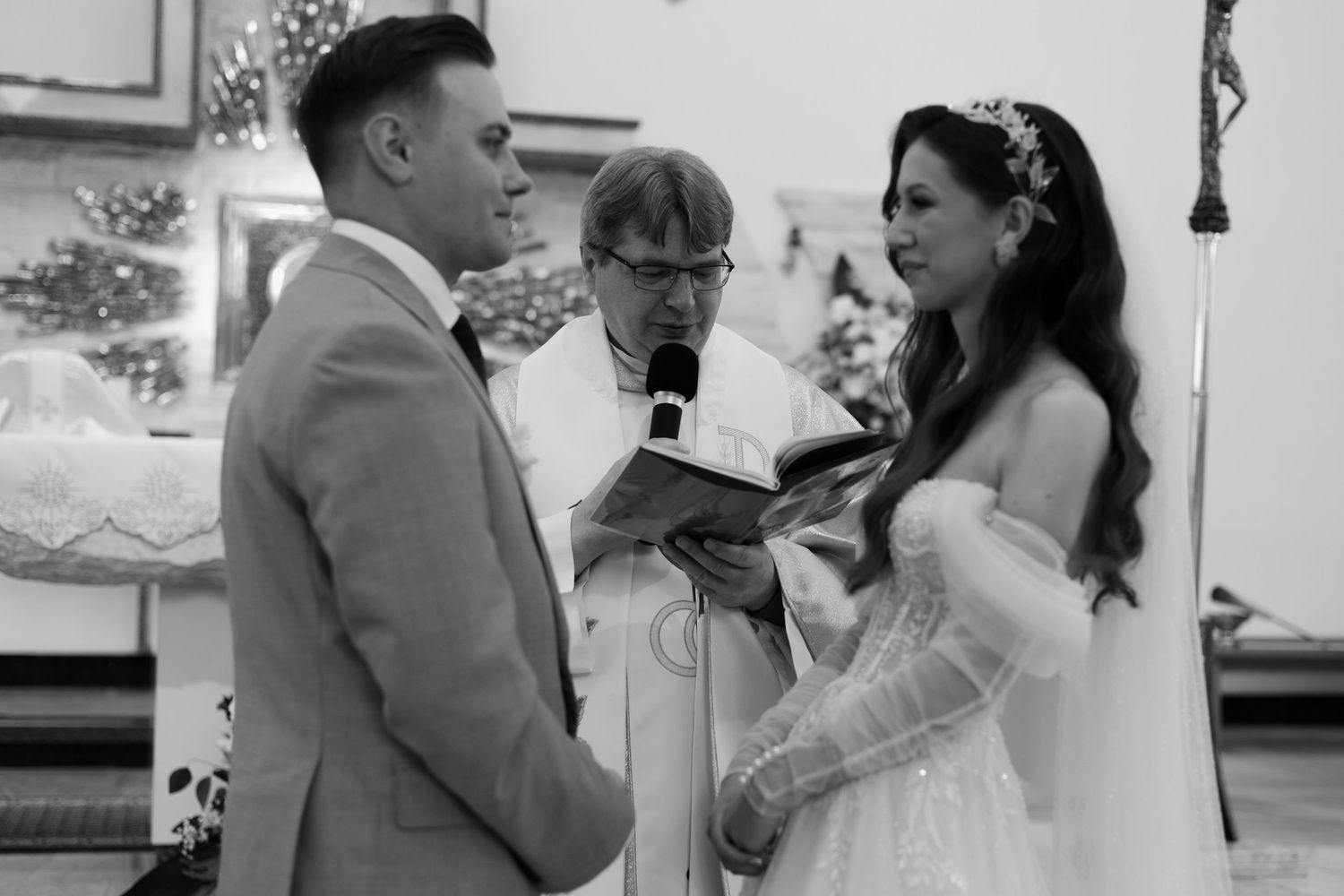 A black and white photograph showing a wedding ceremony with a bride, groom, and officiant in a traditional indoor setting.