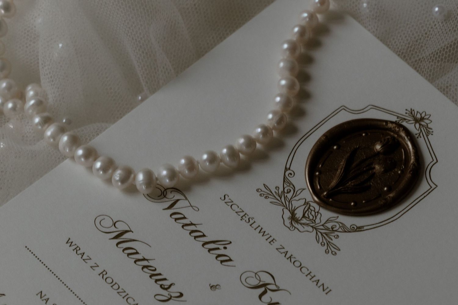 A pearl necklace rests beside an elegant wedding invitation with an ornate vintage crest design.