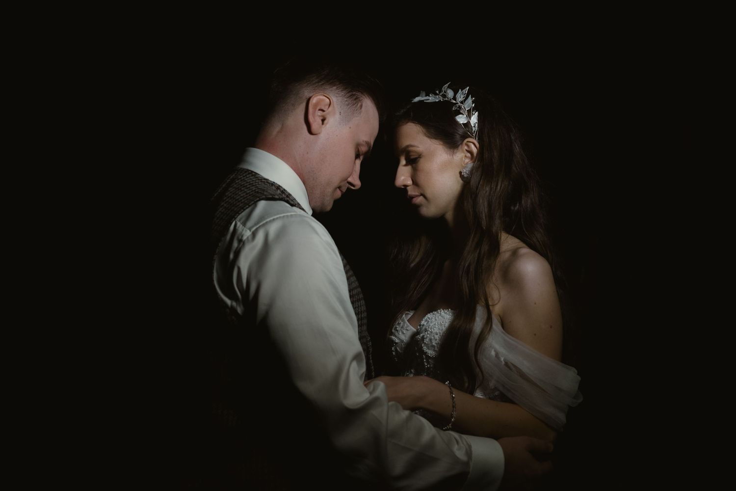 A romantic wedding couple shares an intimate moment in dramatic low-key lighting against a dark background.