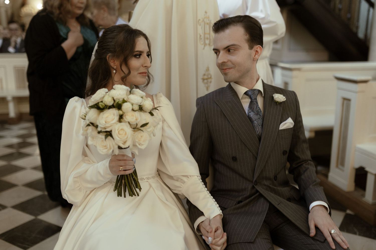 Classic vintage-inspired wedding photo with bride in long-sleeved gown holding white roses and groom in brown suit.
