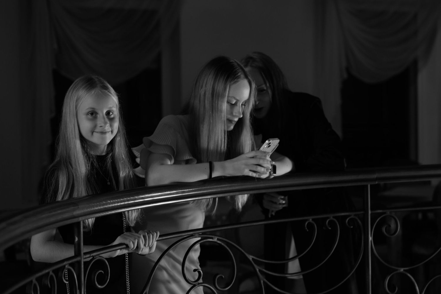Three people leaning on an ornate railing while looking down at a phone in black and white.