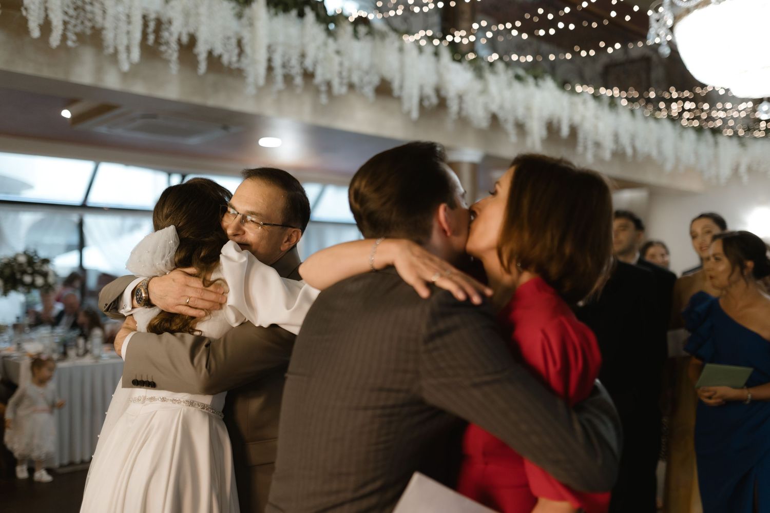 Couples embrace on a dance floor under decorative white hanging decor and string lights at an indoor celebration.