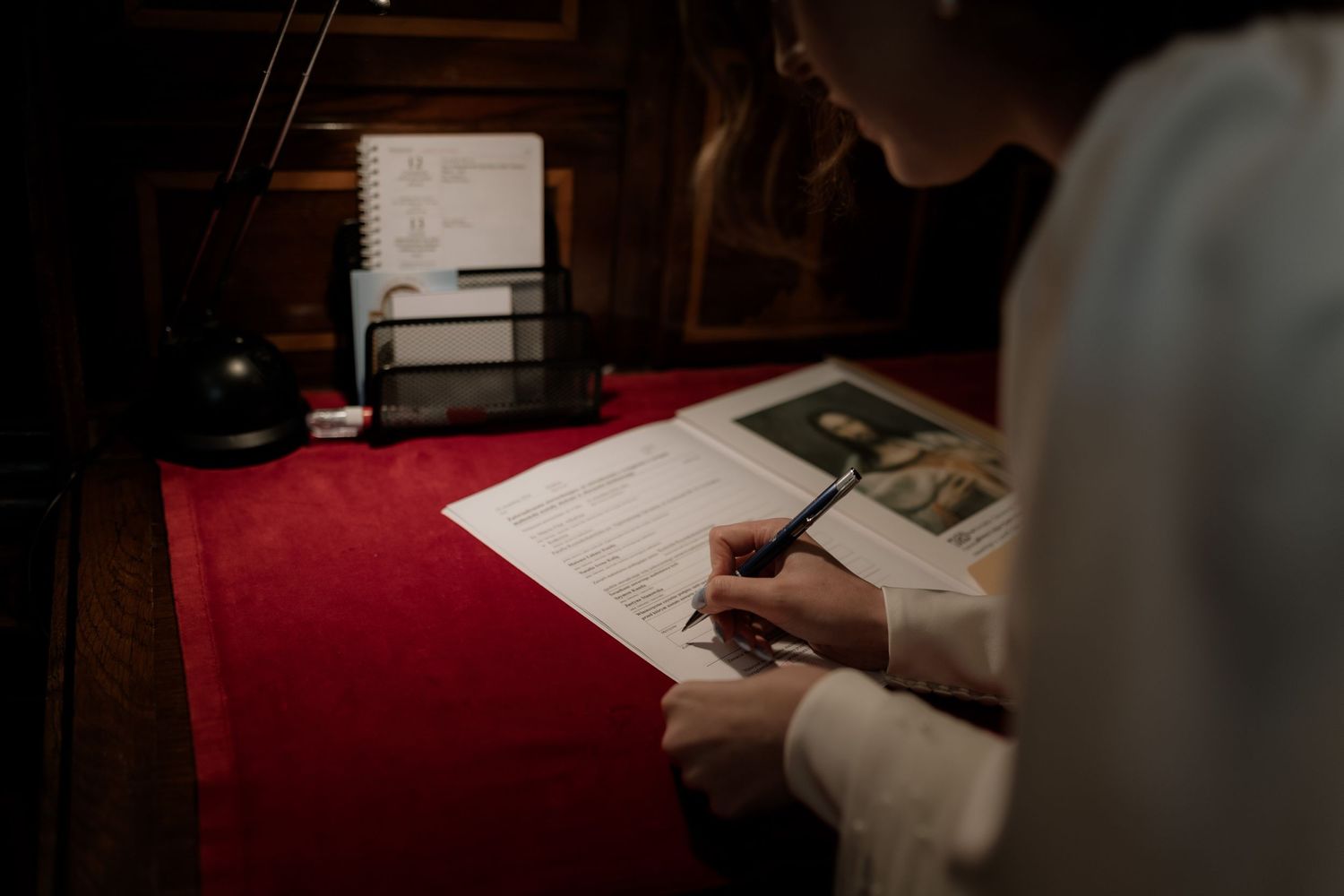 Someone writes with pen in a dimly lit room with documents and a photograph on a red desk surface.