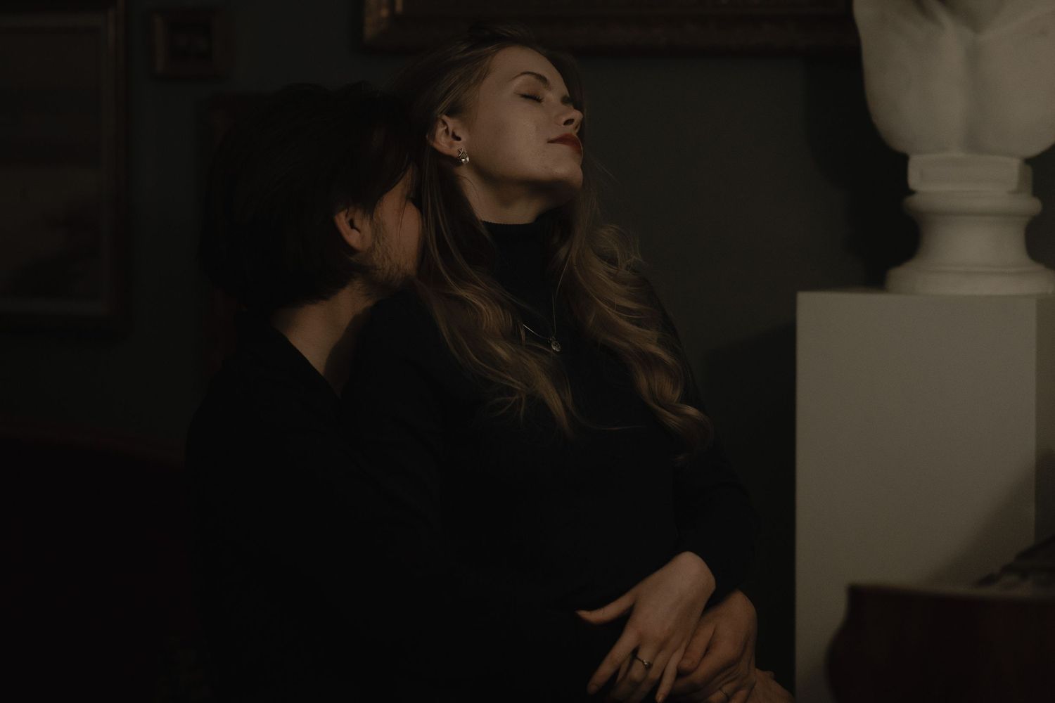 A dark moody portrait of someone in a black outfit resting their head back against a wall in low light.