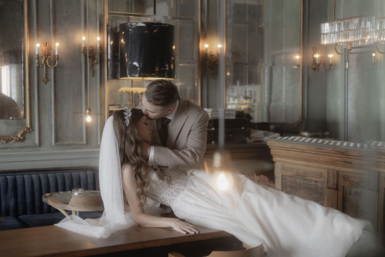 A romantic scene in an elegantly lit room with a couple embracing at a dining table in formal wedding attire.
