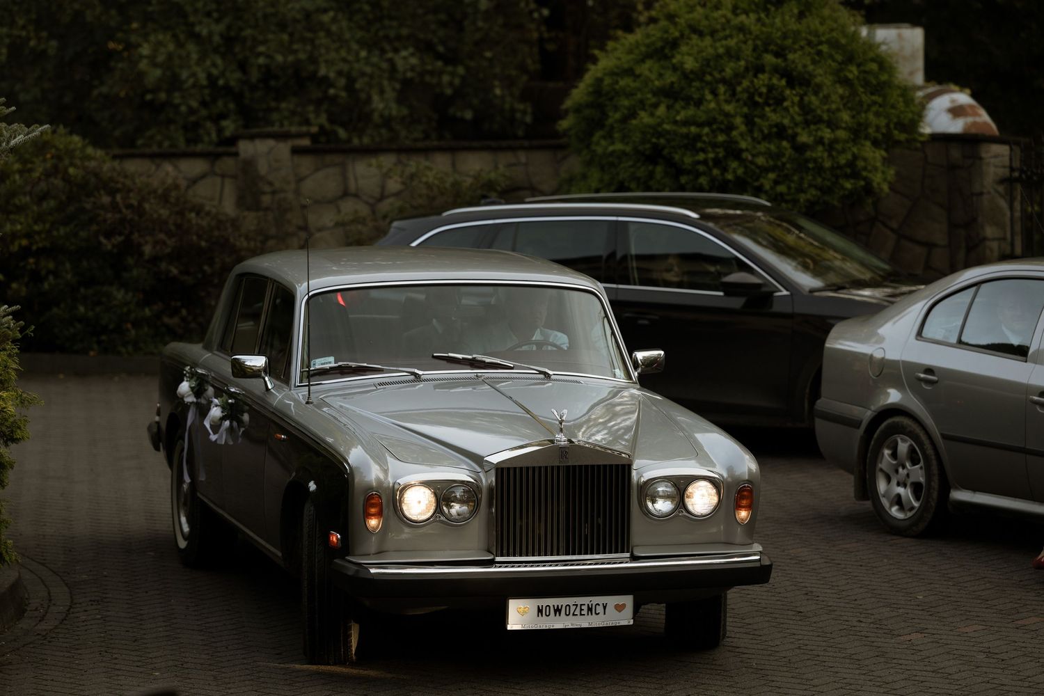 Classic silver Rolls-Royce luxury car parked in a driveway with headlights on at dusk.