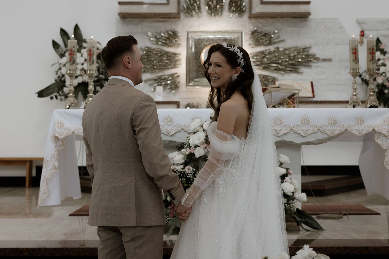 A couple stands at the altar during their wedding ceremony, surrounded by elegant white floral decorations and greenery.
