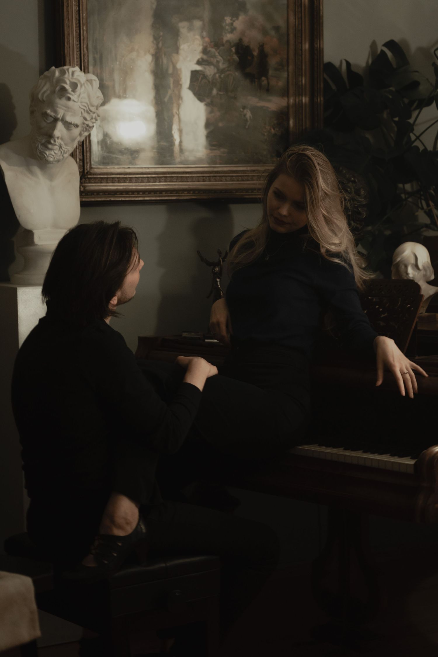 Two people sit in a dark moody room decorated with classical busts and artwork in vintage aesthetic style.