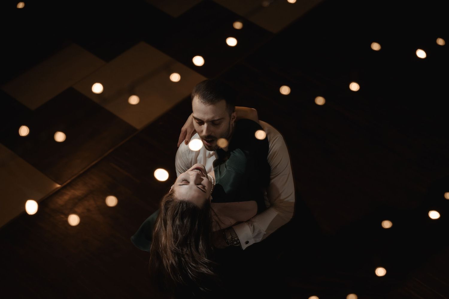 A couple shares an intimate dance under twinkling overhead string lights in a dark room.