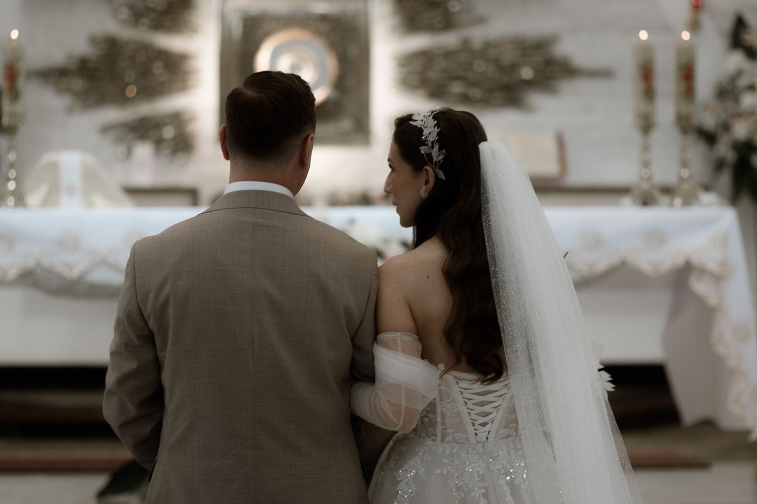 A bride and groom stand at the altar in a church during their wedding ceremony, seen from behind.