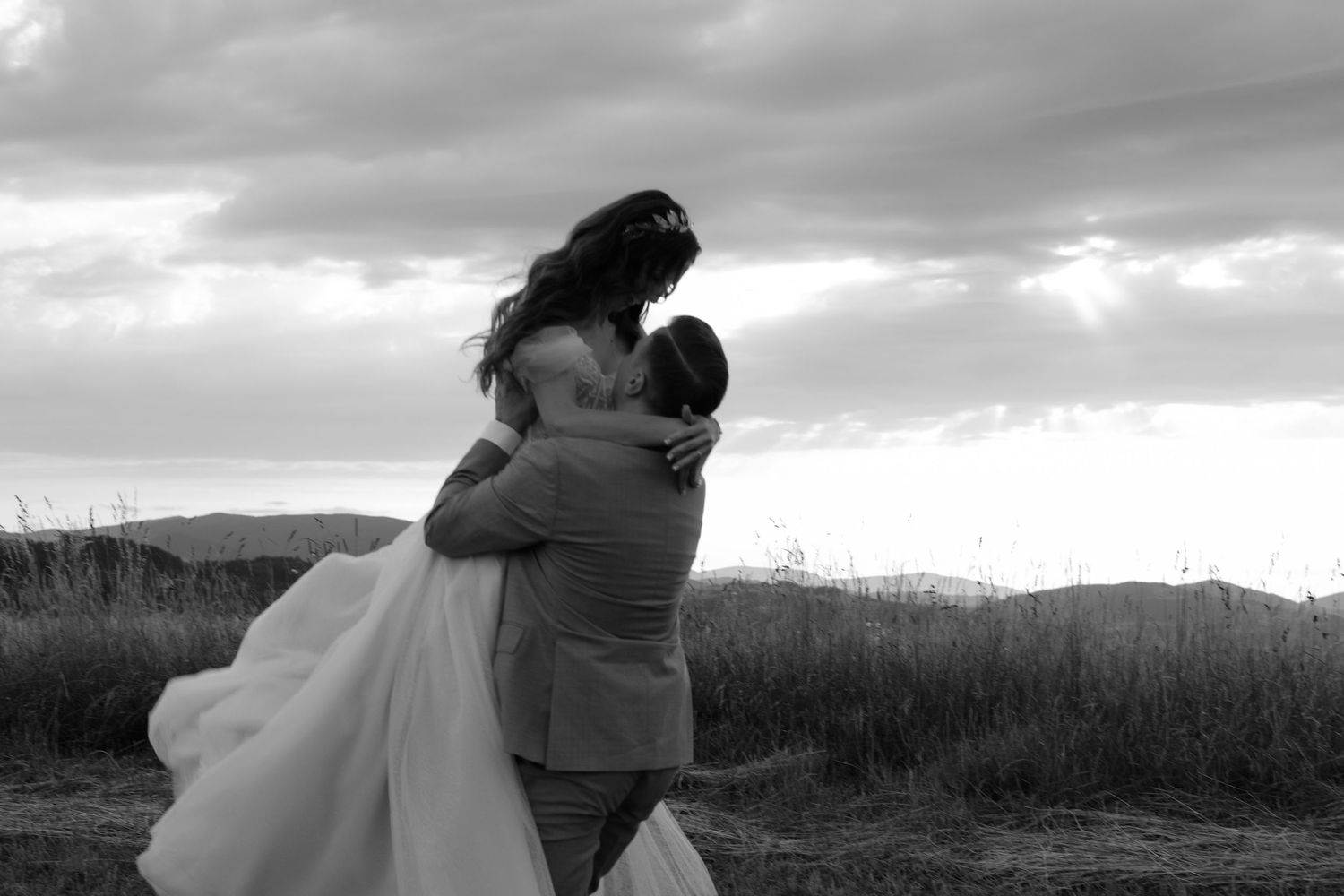 A romantic black and white moment between newlyweds embracing on a hilltop against a dramatic cloudy sky.