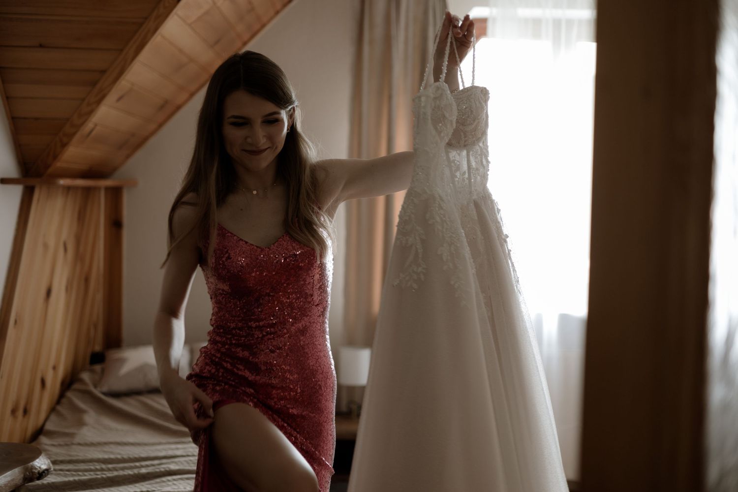 A red satin slip dress hangs in a rustic wooden attic room next to a white wedding gown.